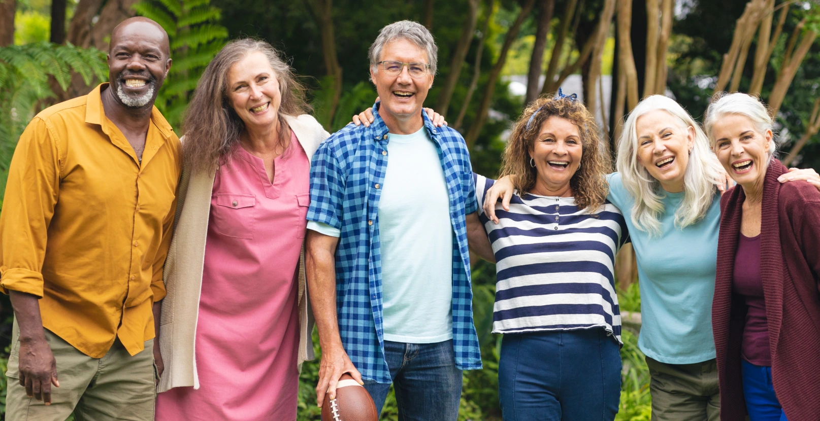 Group of six smiling people standing together outdoors with green foliage background.