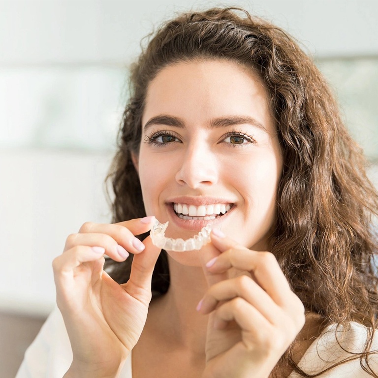 Woman holding clear dental aligners, smiling in bright indoor clinical setting.