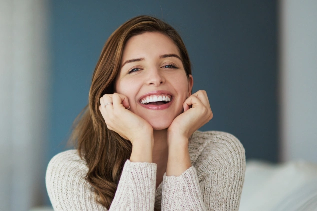 Smiling woman in cream sweater with hands on face, professional dental care setting background.