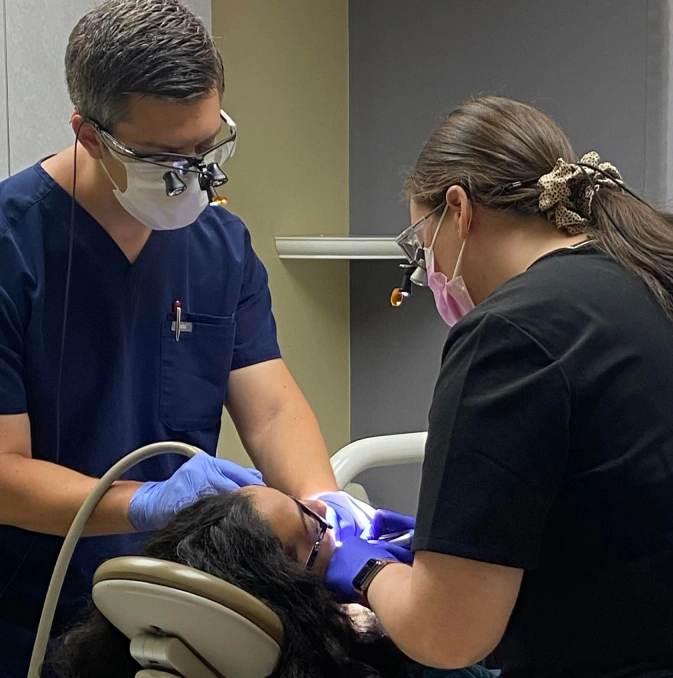 Dentist and assistant performing dental procedure on patient using blue light tool in clinical setting.