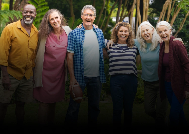Group of six smiling adults standing together outdoors among green foliage and trees.