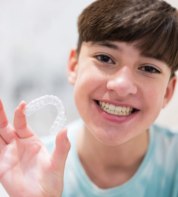 Young boy smiling while holding clear dental aligner or retainer in his hand indoors.
