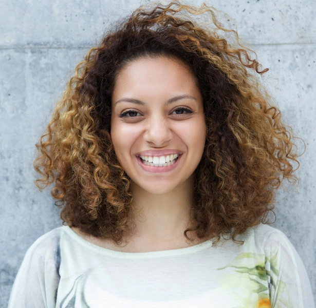 Smiling woman with curly brown hair wearing light colored shirt against gray wall background.