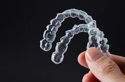 Clear plastic dental aligner held between fingers against dark background