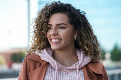 Smiling woman with curly hair wearing brown jacket outdoors in dental care setting.