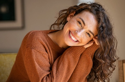 Smiling woman in brown sweater relaxing on couch in warm interior setting.