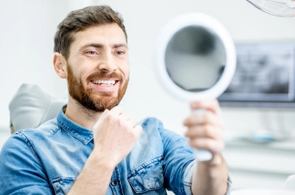 Man smiling while holding dental mirror in bright dental office setting.