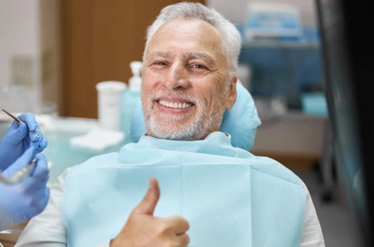 Smiling senior man giving thumbs up in dental chair wearing bib at dentist office.