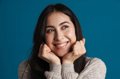 Smiling woman with dark hair resting chin on hands against blue background.