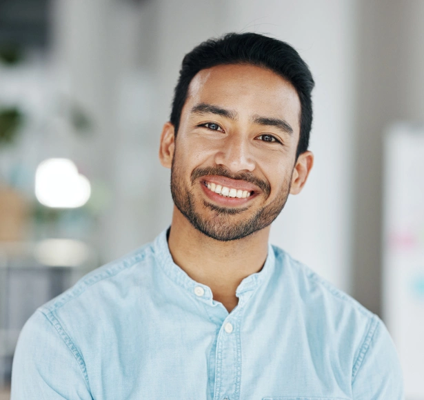 Smiling male dental professional in light blue shirt, indoor clinical setting with blurred background.