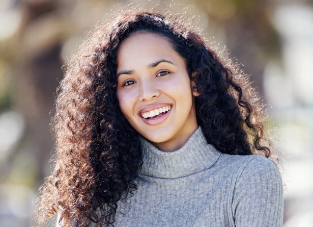Smiling woman with curly hair wearing gray turtleneck outdoors in natural daylight.