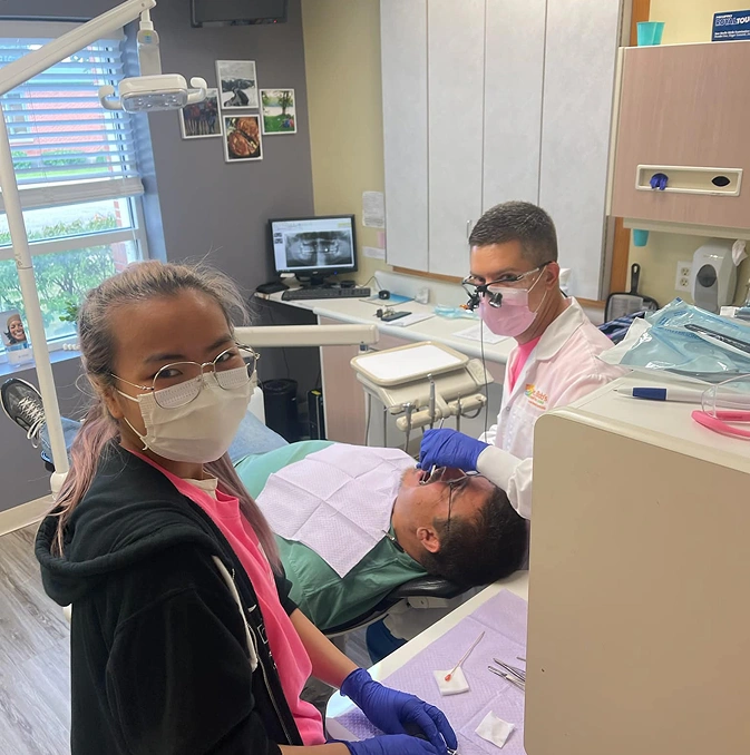 Pediatric dentist examining young patient in dental operatory while assistant observes treatment.