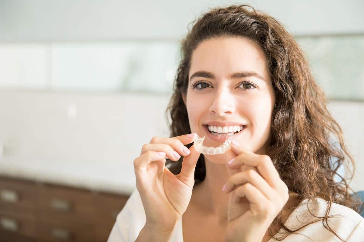Young woman holding clear aligner tray and smiling at camera in dental office.